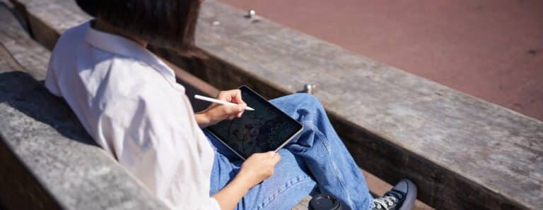 Cropped shot of female hands drawing on digital graphic tablet with pen, sitting with cup of coffee outdoors on bench